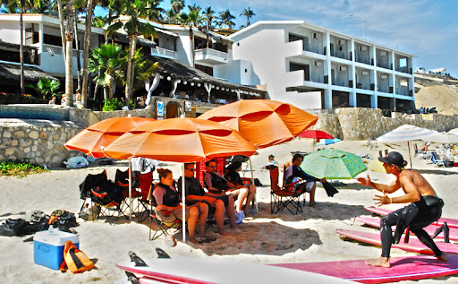 Tourist sitting under the shade of beach umbrellas during a surfing demonstration by High Tide Los Cabos instructor
