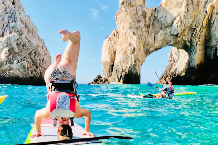 A white person does a headstand on a paddle board near The Arch in Los Cabos. Behind are two other white people on paddleboards. The sky is clear and the sea is shades of blue.