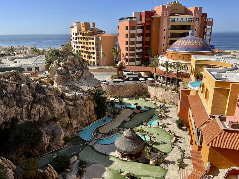 A terracotta-colored hotel in Los Cabos with a pool, blue skies, and a domed structure.