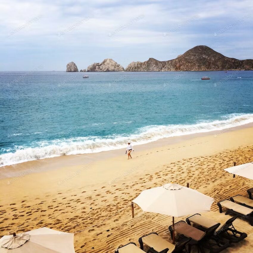Cabo San Lucas Beach with sun loungers, umbrellas, and a cloudy sky. The sea is blue with foamy waves lapping the beach and a mountain in the distance.