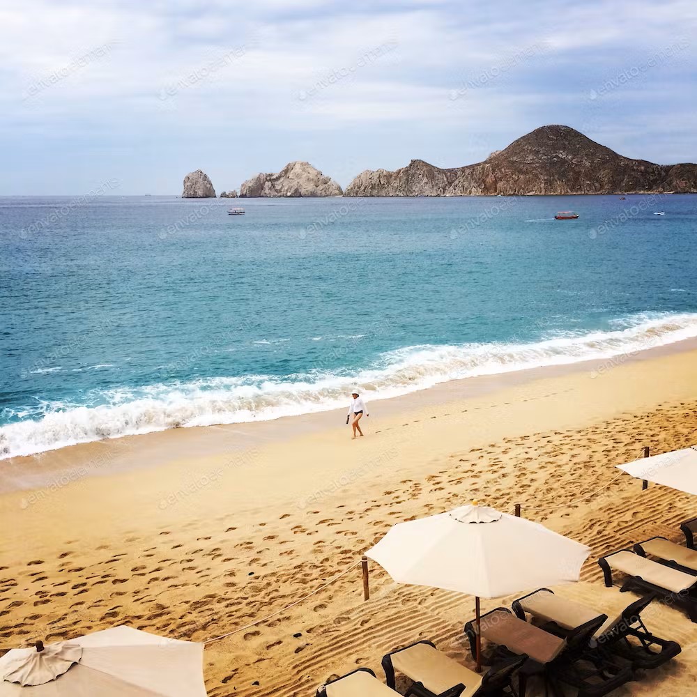 Cabo San Lucas Beach with sun loungers, umbrellas, and a cloudy sky. The sea is blue with foamy waves lapping the beach and a mountain in the distance.